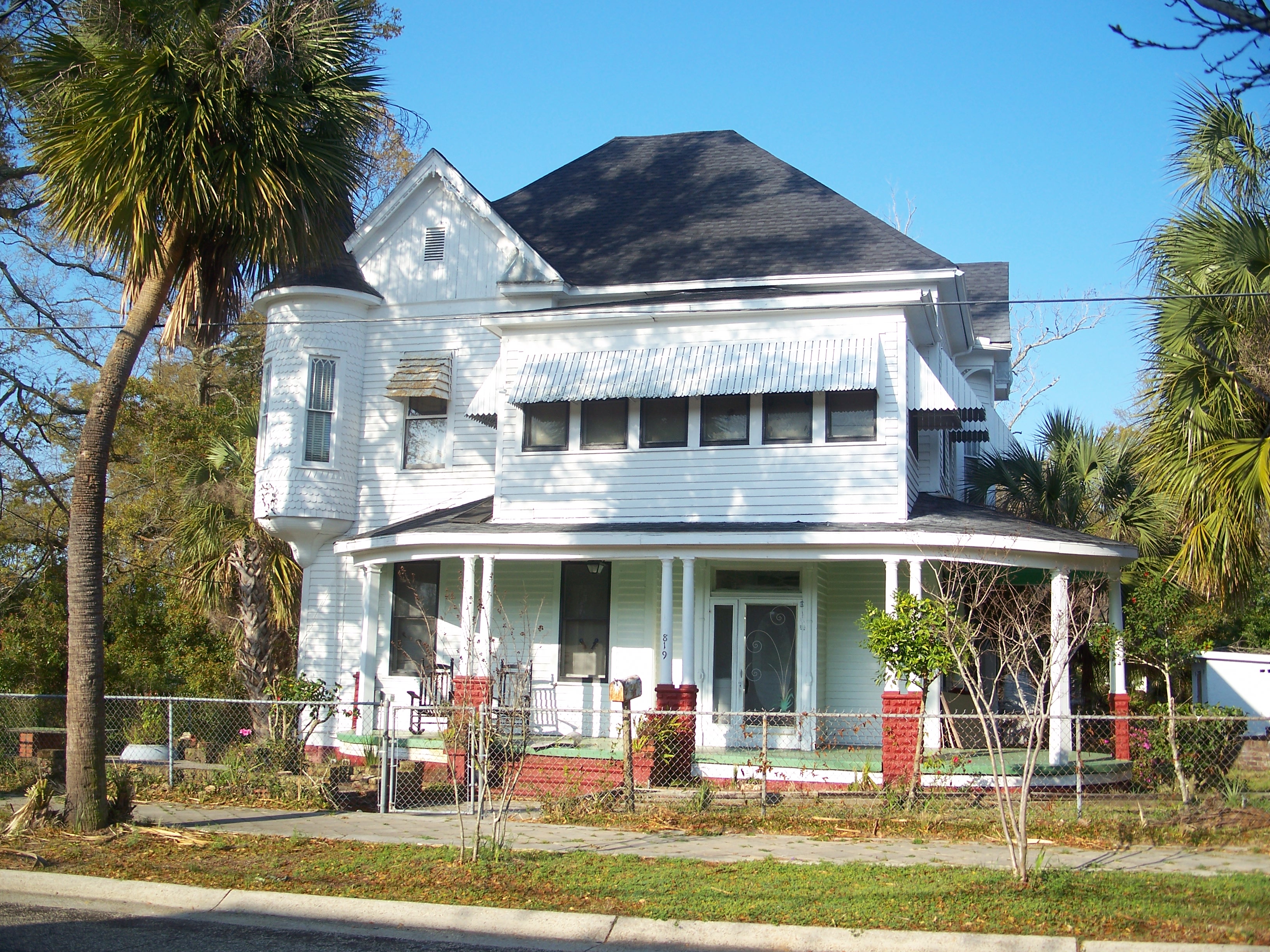 A white two-story wood-frame house with a wraparound porch on a residential street in Pensacola, Florida